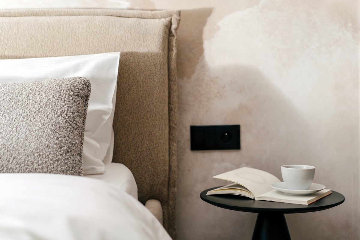 A side table with an open book and a coffee cup beside a made bed at Midis Otel